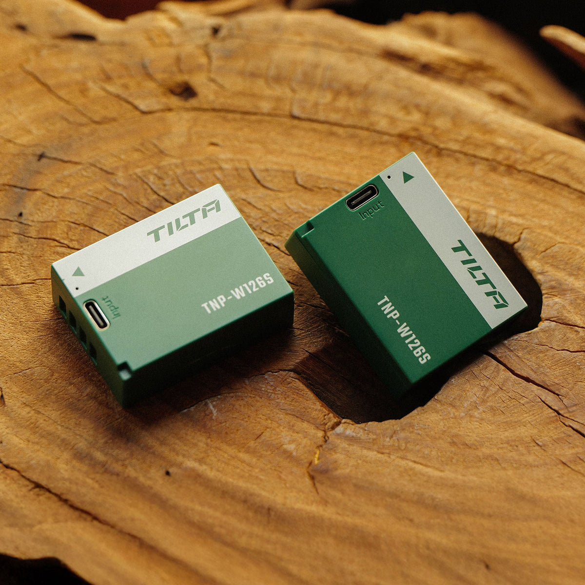 Two Tilta TNP-W126S batteries on a cut tree stump, one Ash Green and one Forest Green, USB-C input ports visible