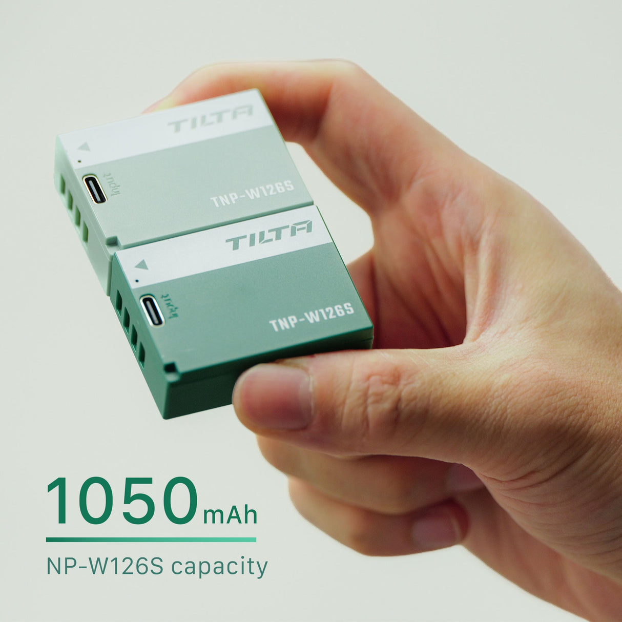 Two Tilta TNP-W126S batteries held between finger and thumb, one Ash Green and one Forest Green, with a 1050mAh NP-W126S capacity callout