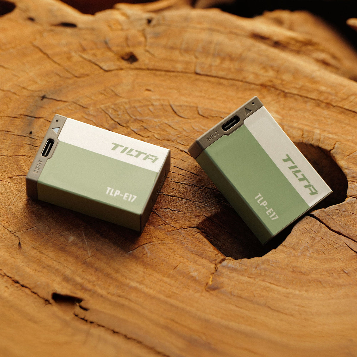 Two Tilta TLP-E17 batteries in Ash Green resting on a cut tree stump with their USB-C input ports facing forward