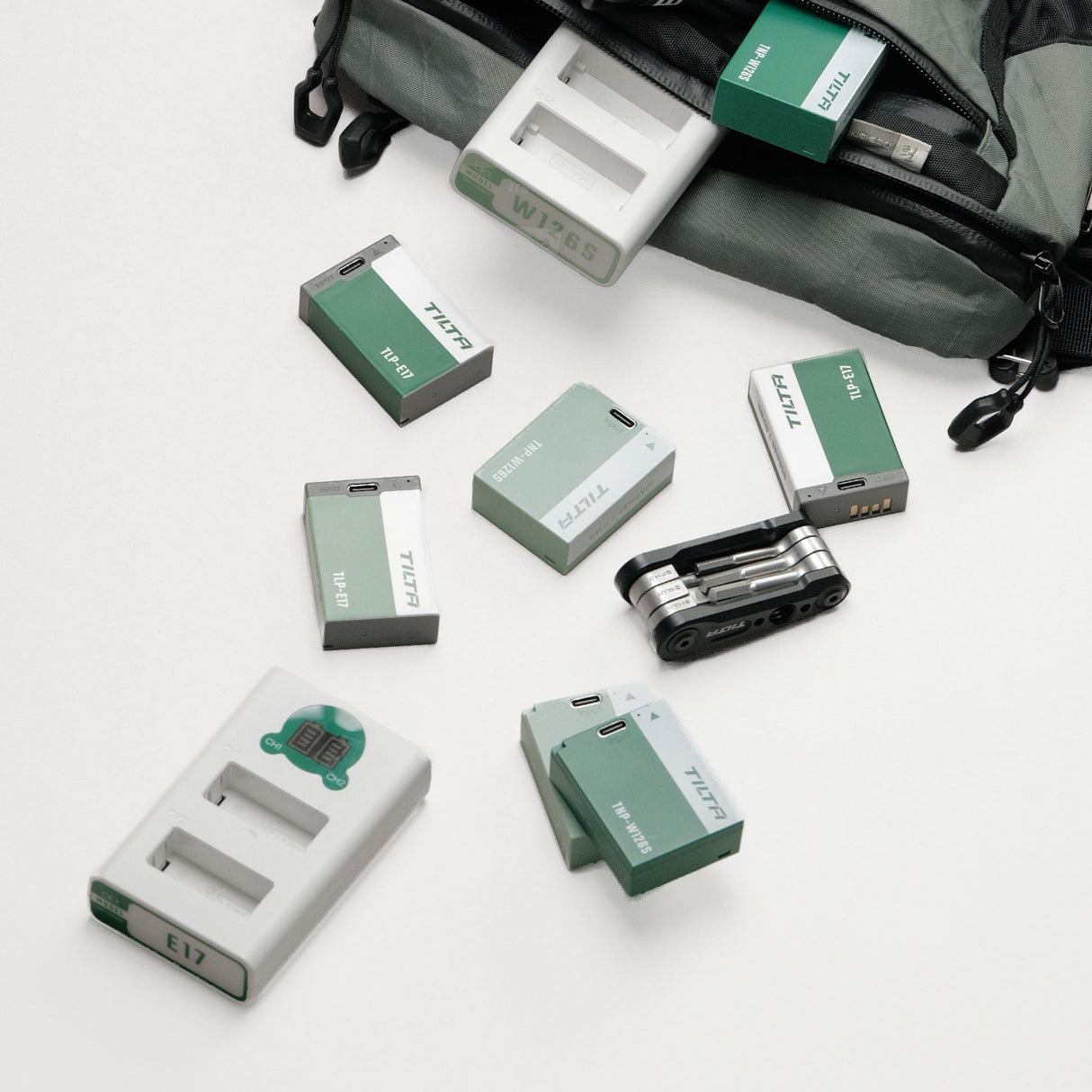 Tilta TBC-E17 LP-E17 dual charger and TLP-E17 batteries in Ash Green and Forest Green laid out beside a camera backpack